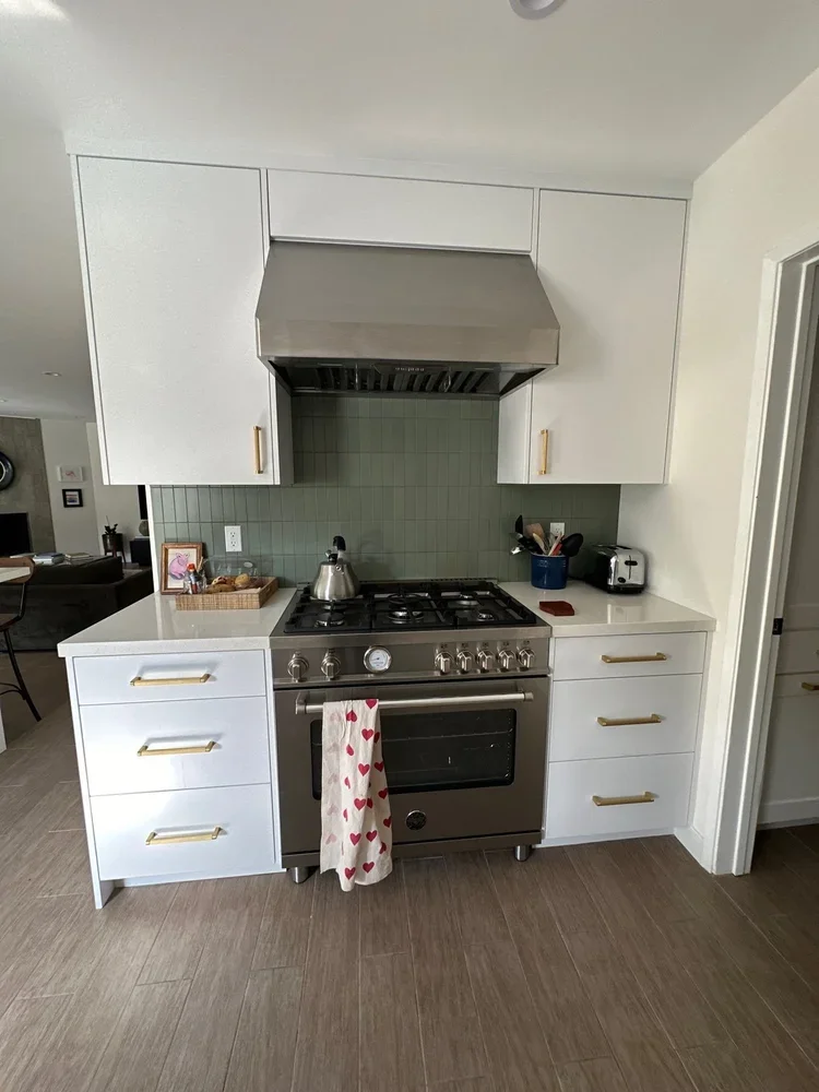 Kitchen with white cabinets, a stainless steel stove and range hood, a teal backsplash, and a towel with red hearts hanging from the oven handle.