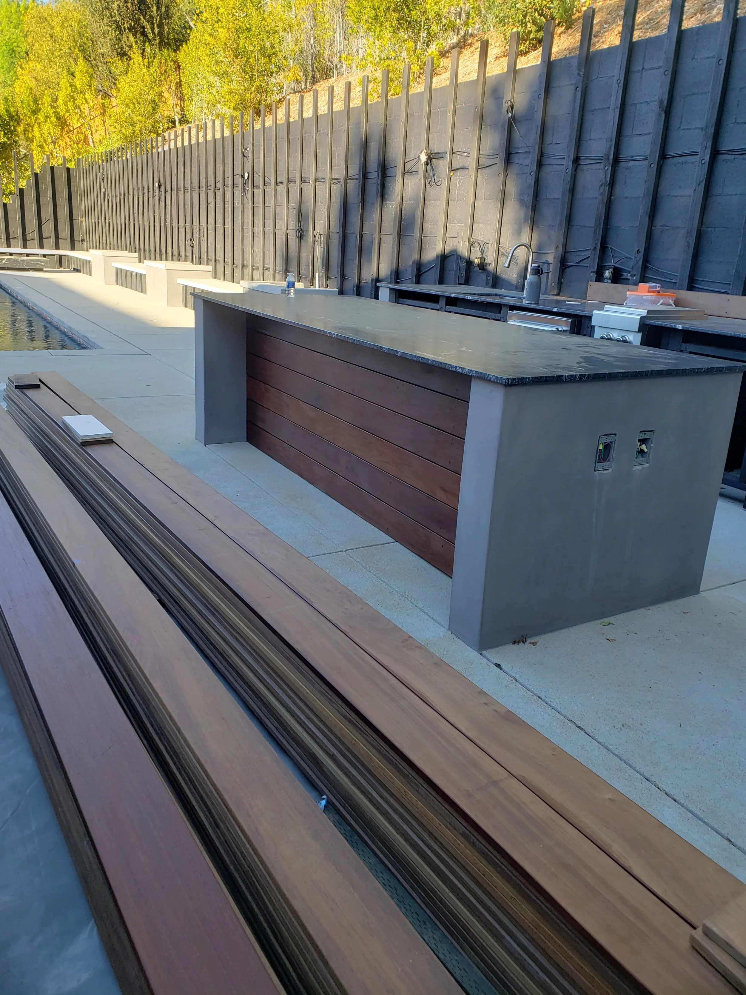 Outdoor kitchen area with concrete counter and sink, construction materials, and a black wooden fence. Trees with green and yellow leaves are in the background.