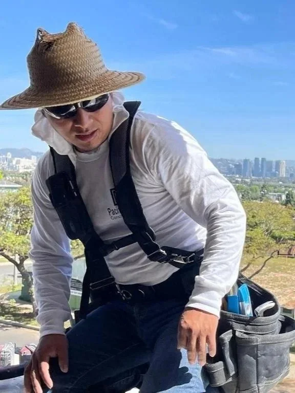A man wearing a straw hat, sunglasses, and a white long-sleeve shirt with a backpack, working on a ladder outdoors with trees and city buildings in the background.