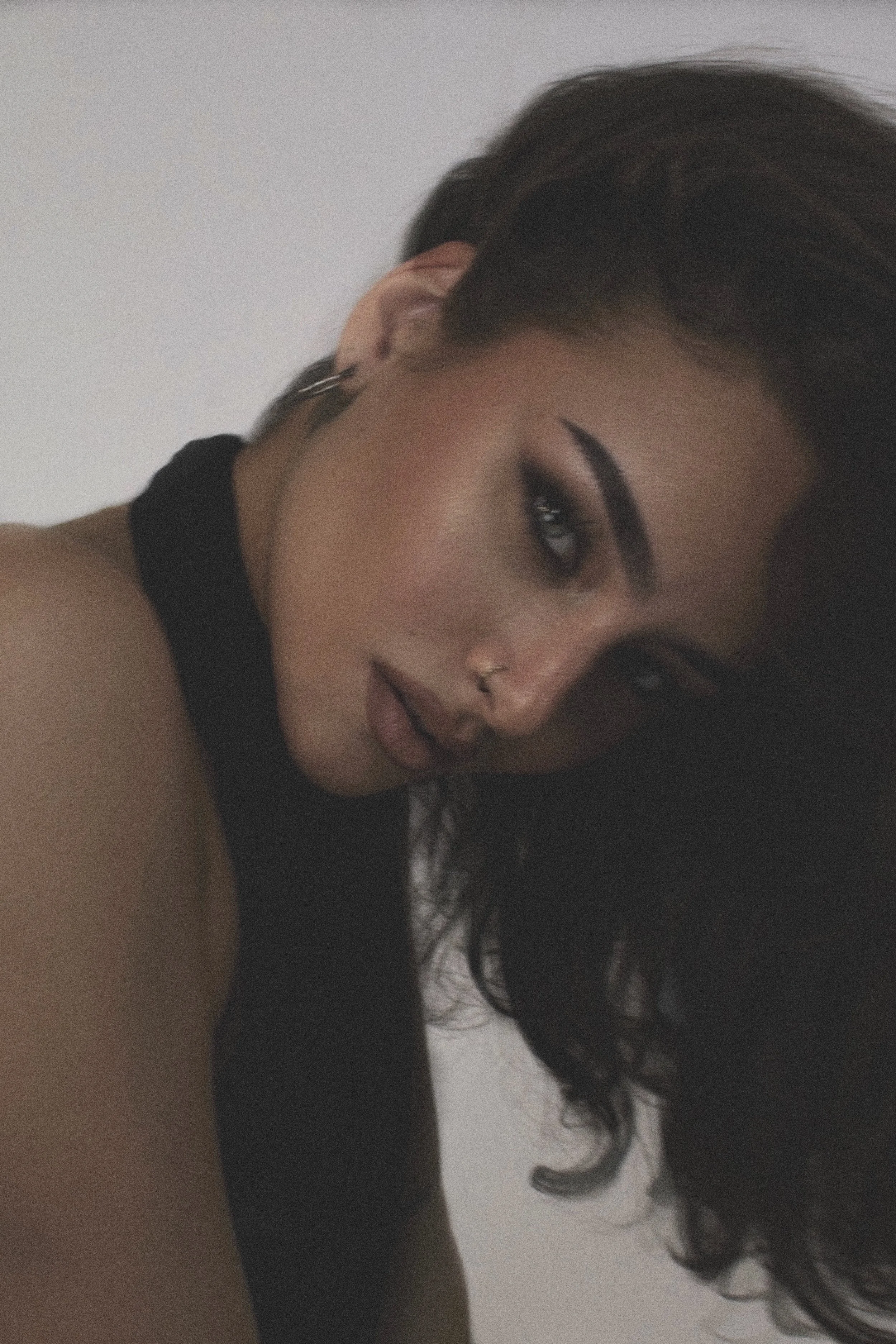 Close-up of a woman with dark hair, makeup, and a nose ring, wearing a black sleeveless top, lying on her side against a plain background.