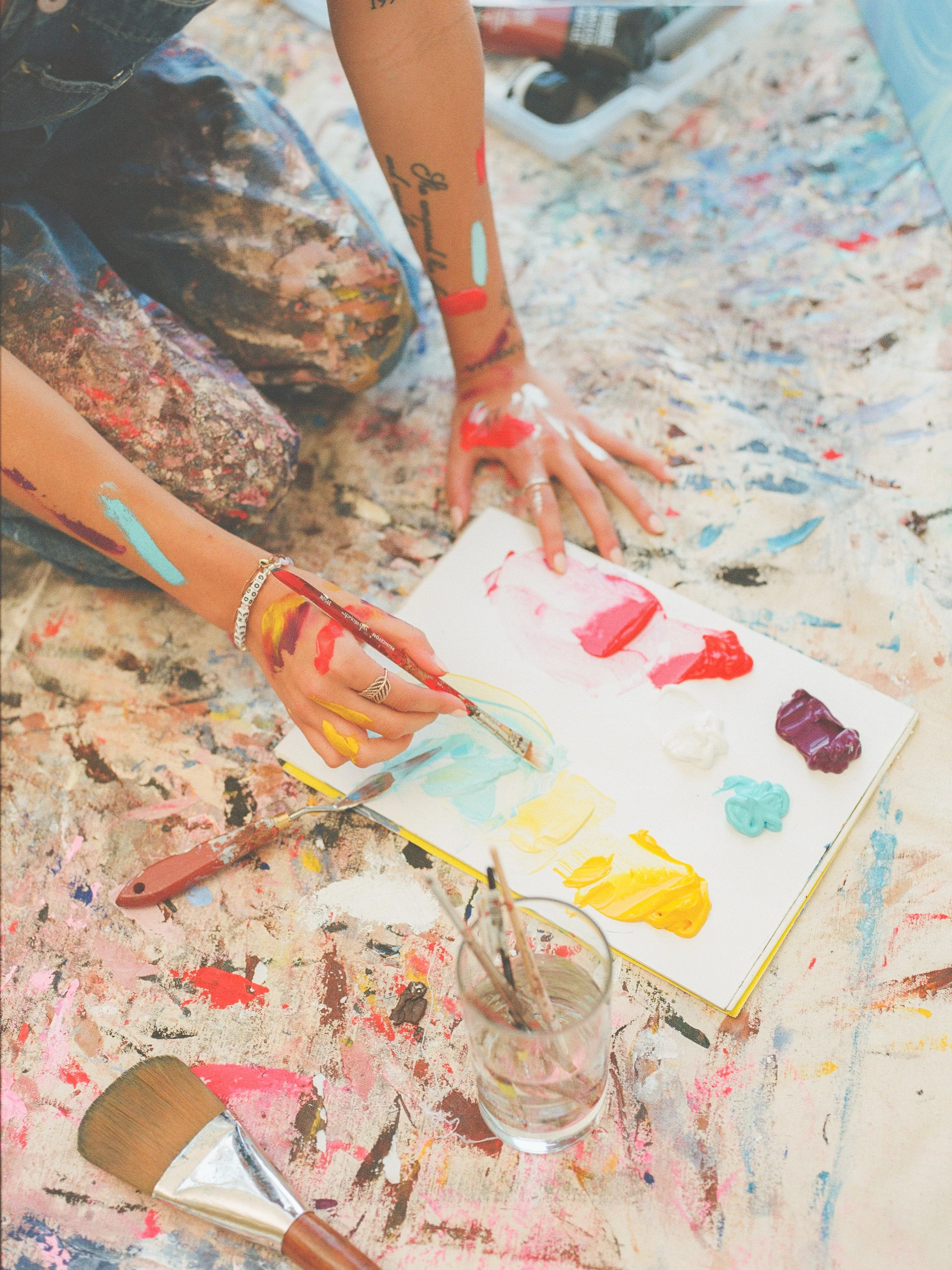A person painting with red, yellow, blue, and purple acrylic paints on paper, surrounded by splattered and dripped paint on a messy table, with paintbrushes and containers.