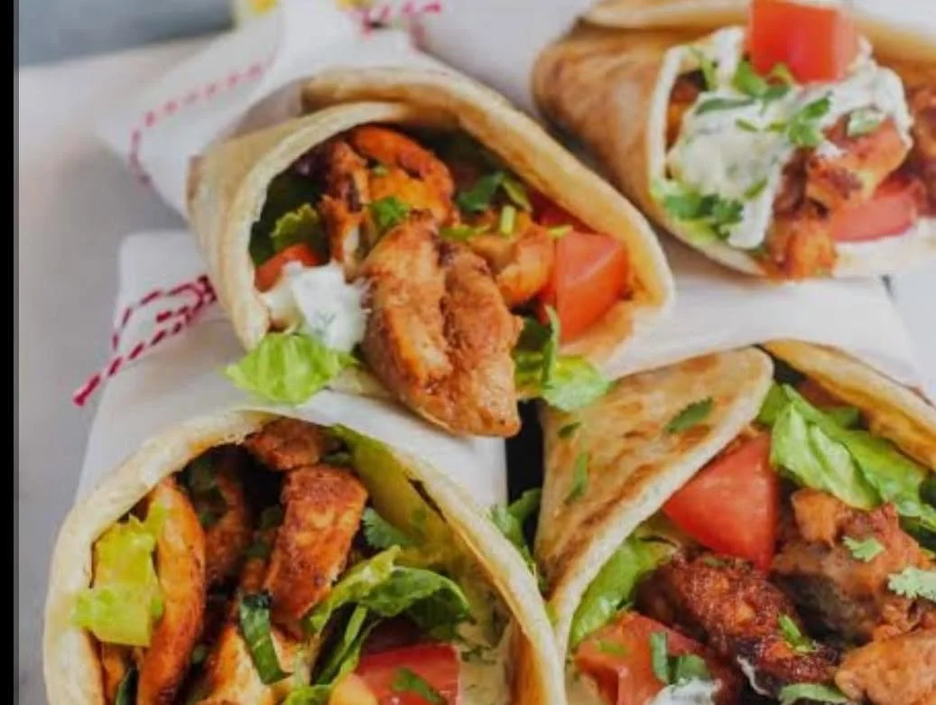 Close-up of four tacos filled with fried chicken, lettuce, tomato, and cilantro on a white surface.