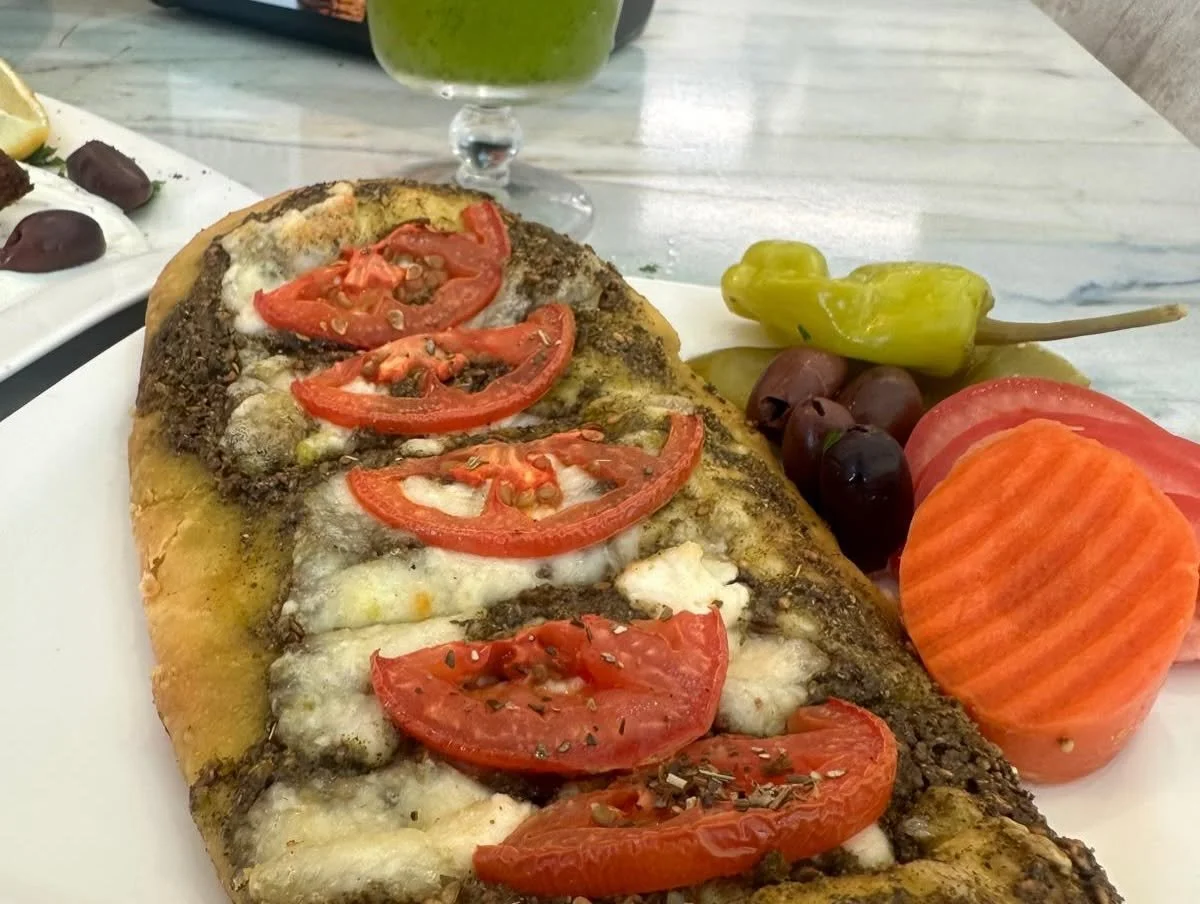 Baked fish fillet topped with tomato slices and herbs, served with pickled peppers, black olives, and sliced tomatoes on a white plate.
