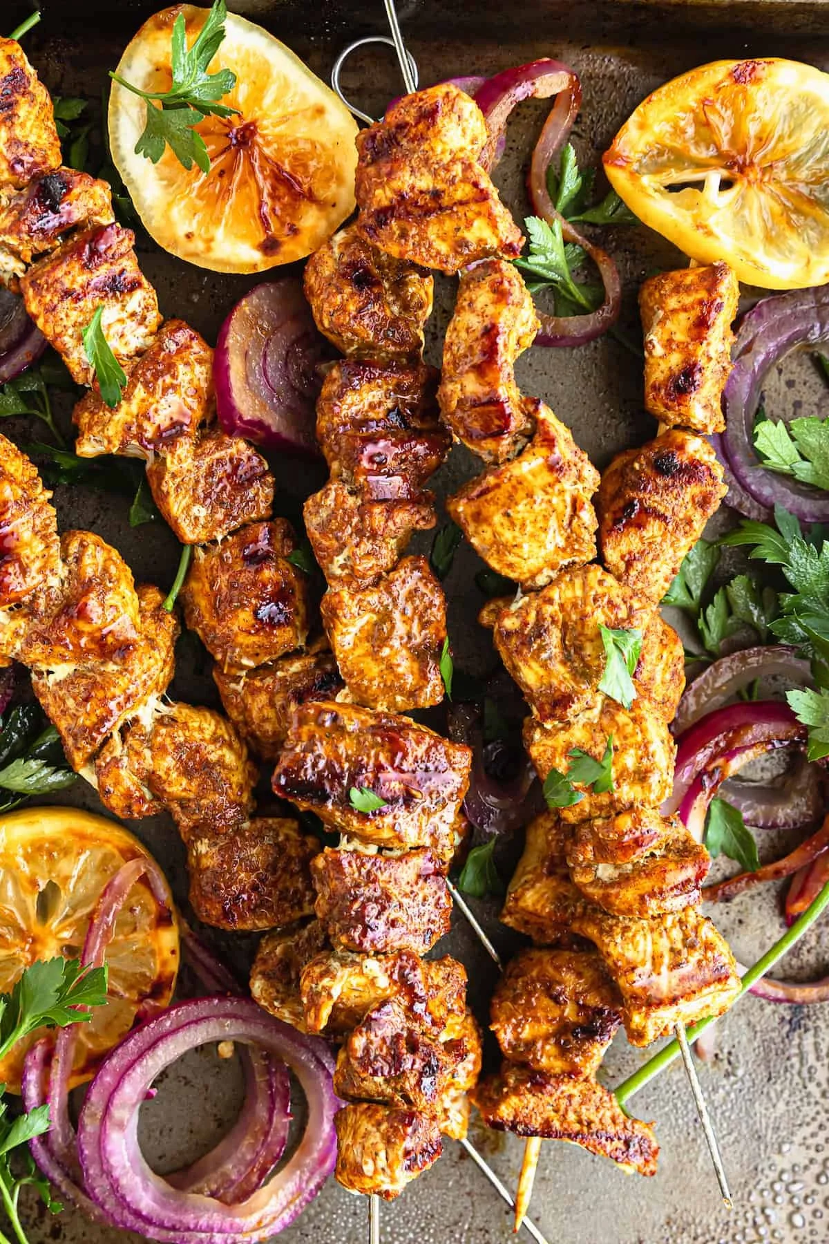 Close-up of grilled chicken skewers garnished with fresh parsley, slices of lemon, grilled tomato halves, and red onion rings, on a rustic surface.