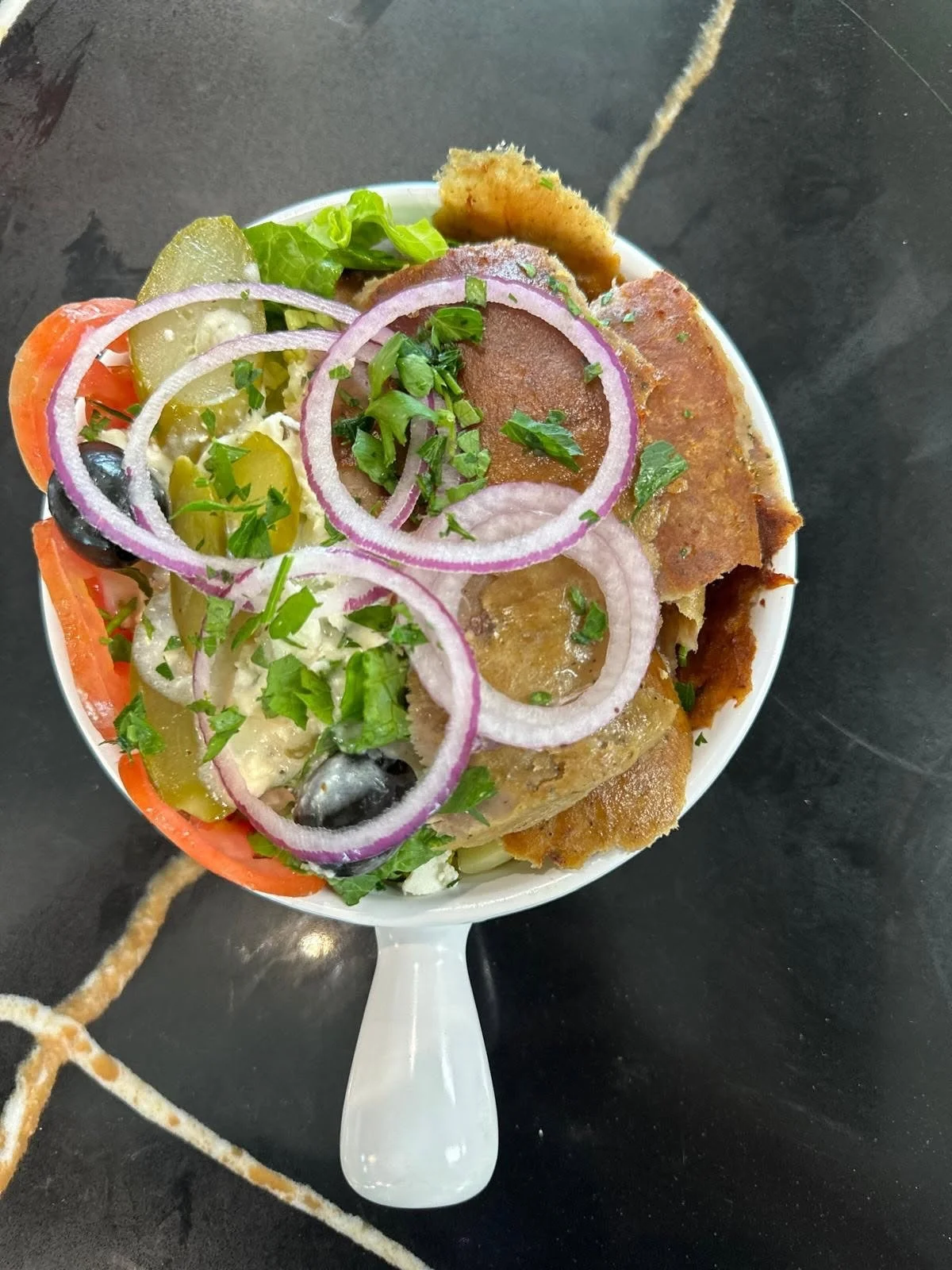 A bowl of salad with sliced onions, chopped green herbs, tomatoes, pickles, black olives, and pieces of fried food garnished with herbs, served in a white bowl on a black surface.