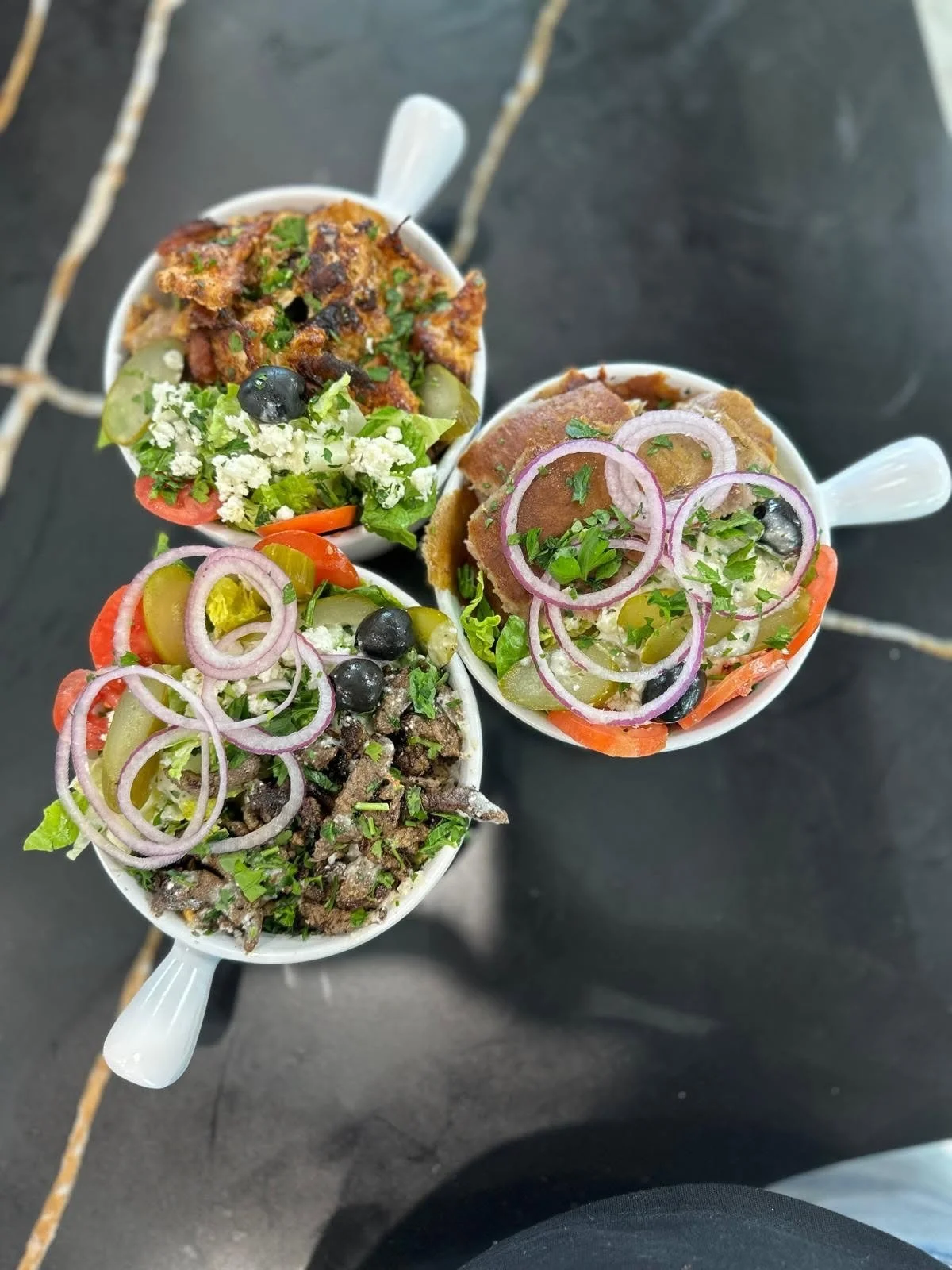 Three small white cups filled with colorful salads and toppings, including lettuce, tomatoes, onions, olives, and various meats, on a black surface.