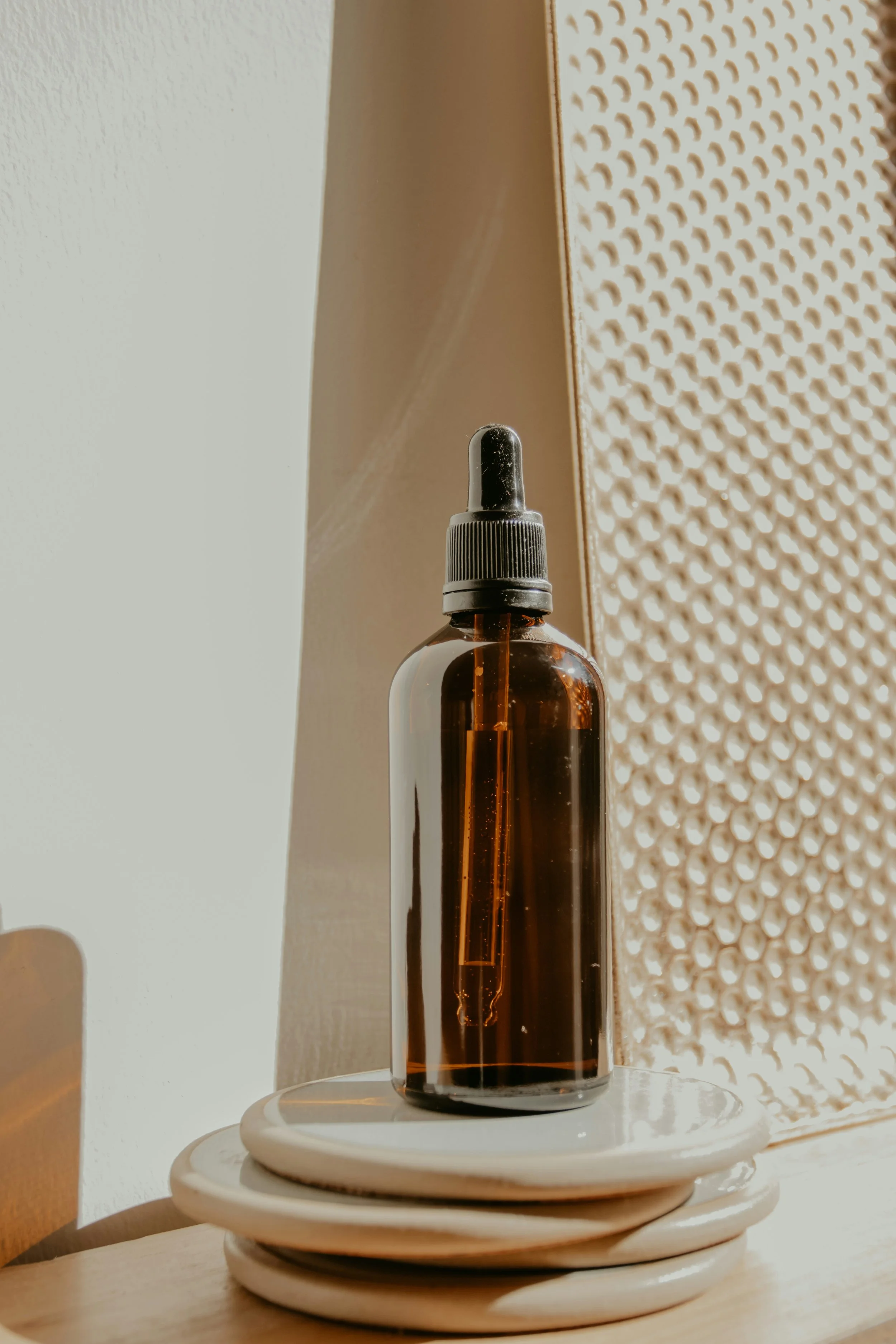 A brown glass dropper bottle placed on a stack of white plates, with warm sunlight casting shadows and a textured wall in the background.