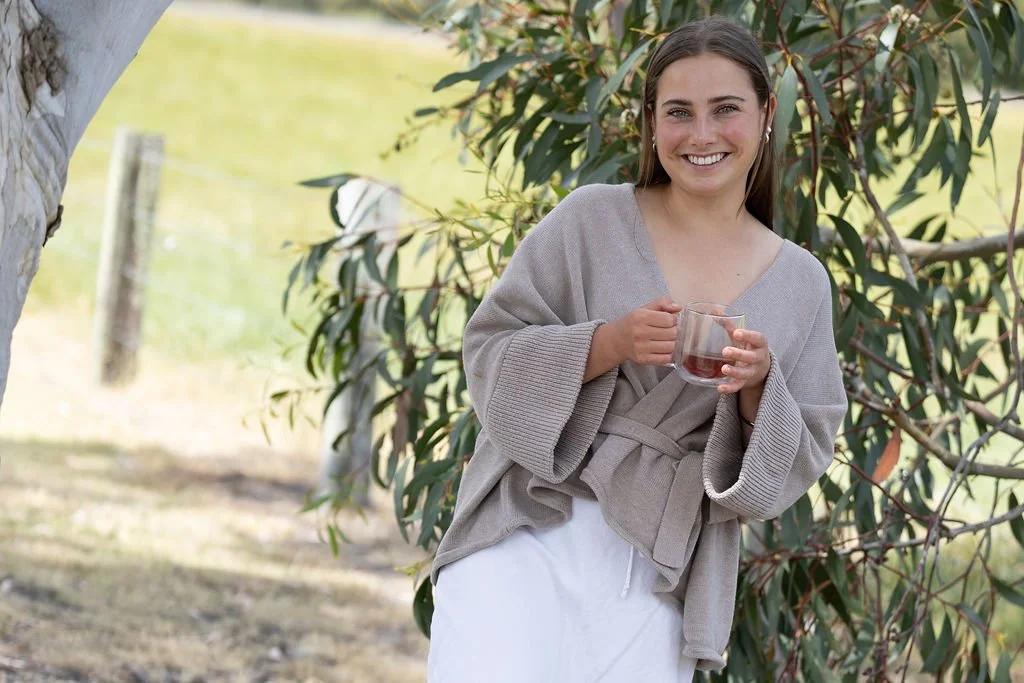A woman standing outdoors near a tree, holding a glass cup of coffee or tea, smiling, dressed in a light gray cardigan and white skirt.