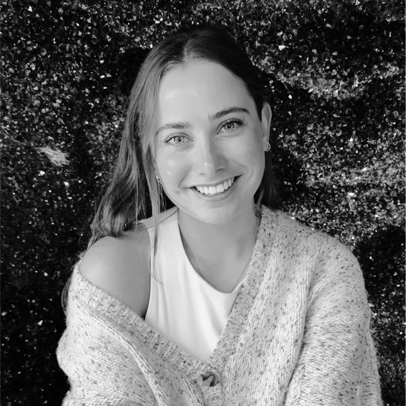 Black and white photo of a young woman with long hair, smiling, wearing a light-colored sleeveless top and a cozy knit sweater, standing in front of a dark, glittery background.