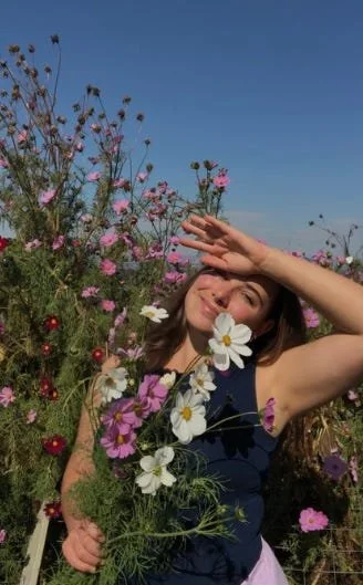 Young woman smiling amidst colorful wildflowers in a field, shielding her eyes from the sun with her hand against a clear blue sky.