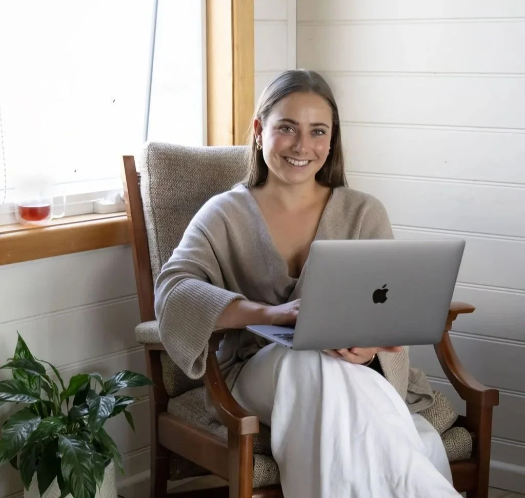 A young woman with long hair, smiling, sitting in a wooden chair with light upholstery, using an open MacBook on her lap in a cozy room with white shiplap walls, a large window, a plant in a white pot, and a drink on the windowsill.