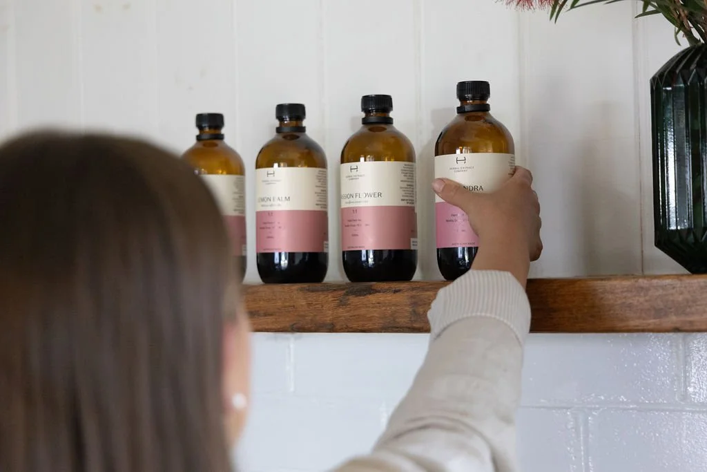 A person with long hair reaches for a brown bottle labeled 'Hydration' on a wooden shelf, with three other similar bottles behind it in a row, in a room with a white wall and a plant on the right.