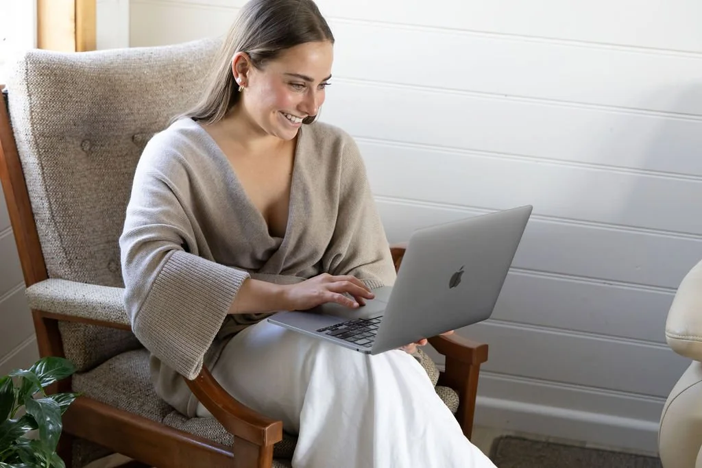 A woman sitting in a comfortable chair, smiling while using a laptop in a cozy, well-lit room.