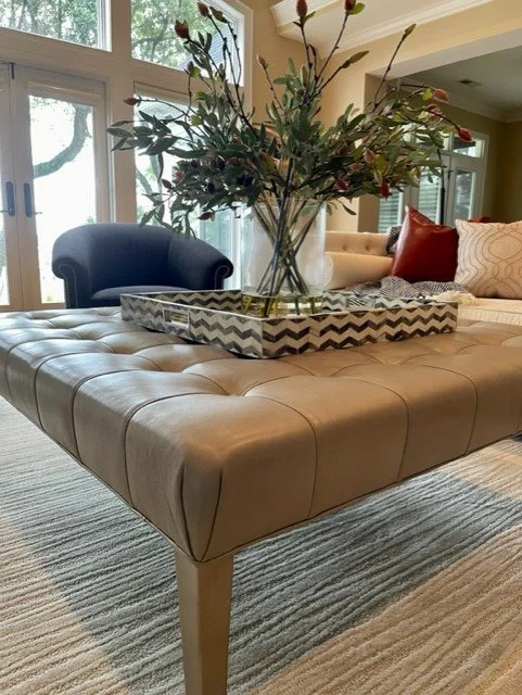 A tufted beige leather ottoman with a chevron-patterned tray holding a large glass vase filled with an arrangement of flowering branches, in a living room with large windows and seating including a navy armchair and a beige sofa with red and patterned pillows.