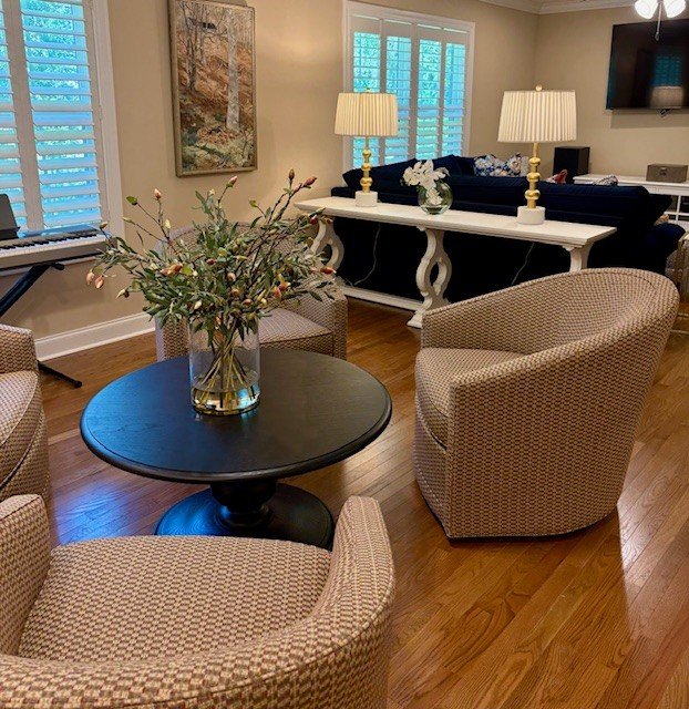 Living room with floral arrangement on a black round table, surrounded by beige upholstered chairs, a sideboard with lamps, a black sofa with pillows, a piano, and a wall-mounted television, with hardwood floors and windows with shutters.