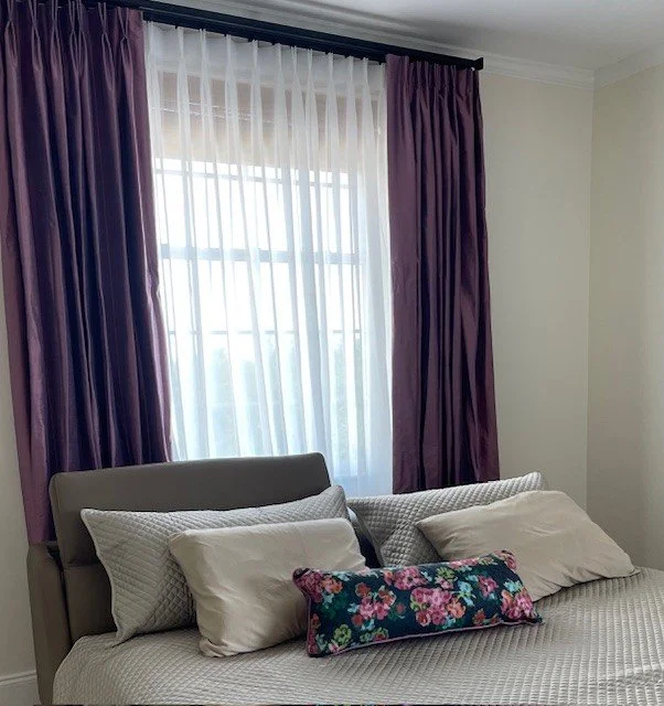 A bedroom with purple curtains, a gray headboard, and a bed with white sheets, three white pillows, and a floral decorative pillow.