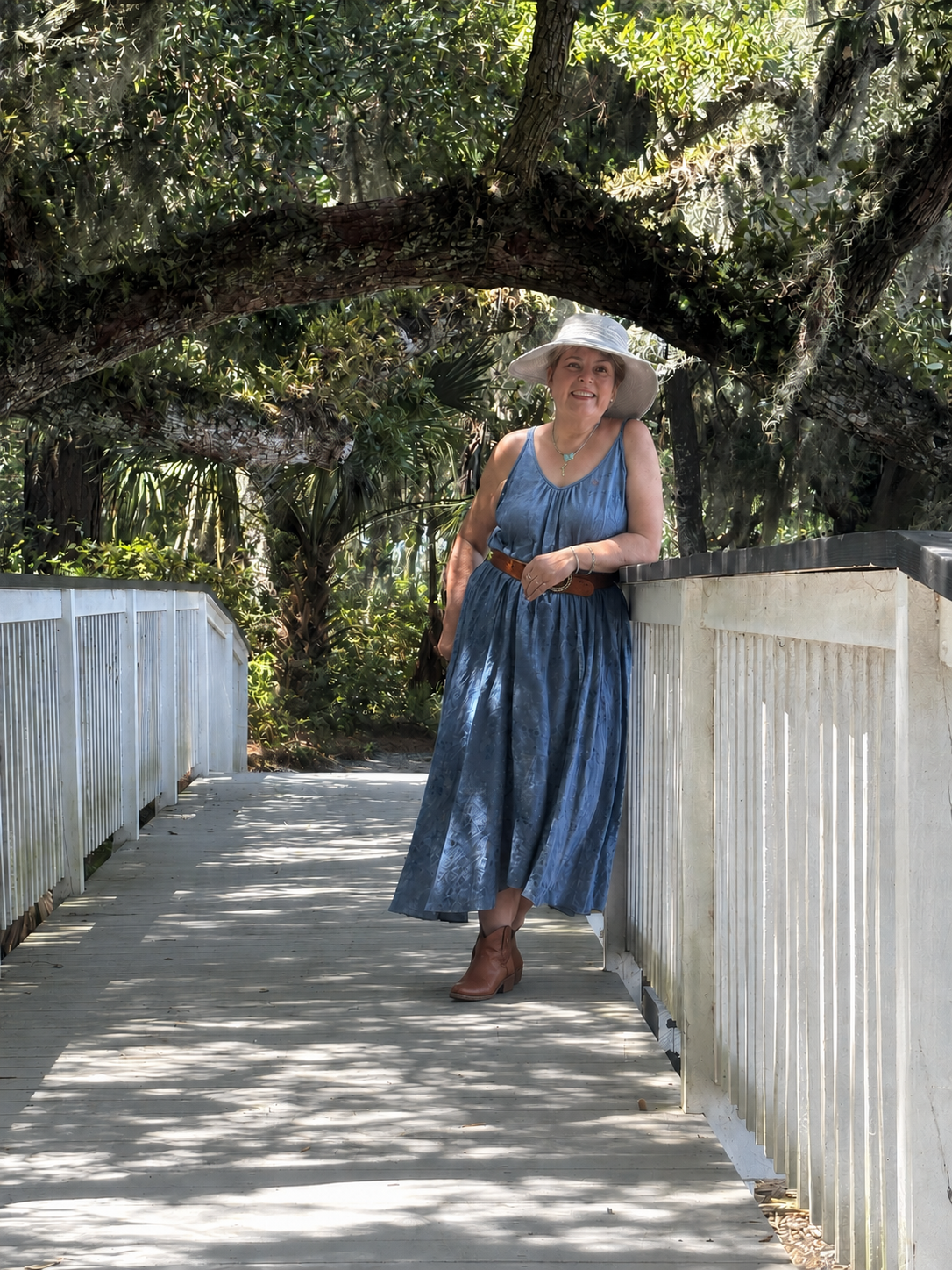A woman wearing a blue dress, tan hat, and brown ankle boots standing on a wooden walkway amidst lush greenery and large trees, smiling at the camera.