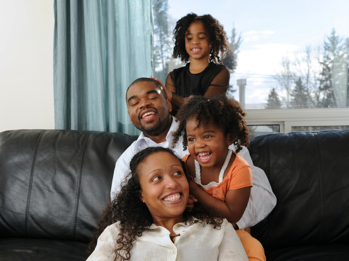 A happy family of four, including a man, a woman, a young girl, and a toddler girl, sitting and playing together on a black leather couch in a living room with a large window showing trees outside.