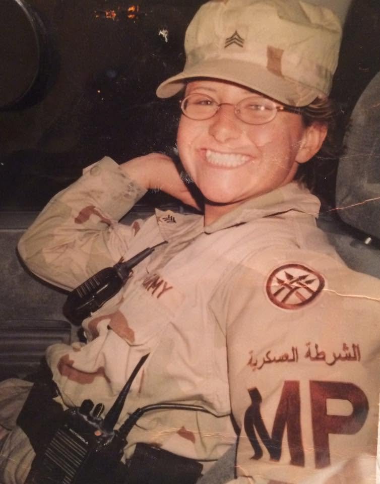 A smiling woman in a military uniform with a cap, glasses, and a beige camouflage pattern, sitting inside a military vehicle.