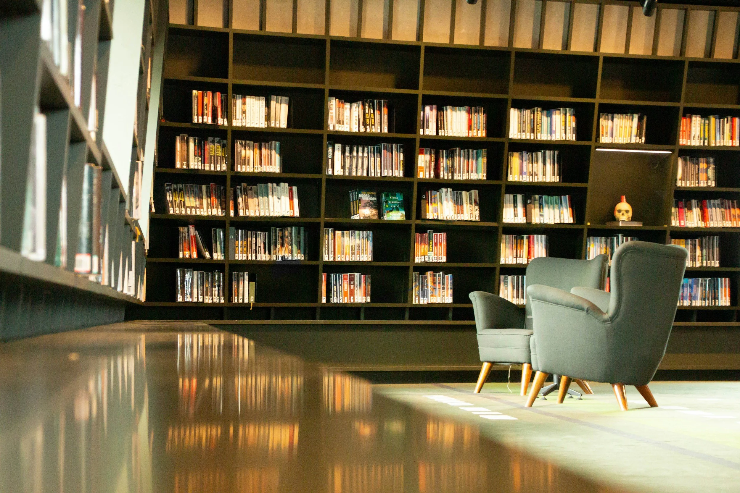A bookshelf filled with books in a library setting, with two gray armchairs in the foreground and a reflective wooden floor.