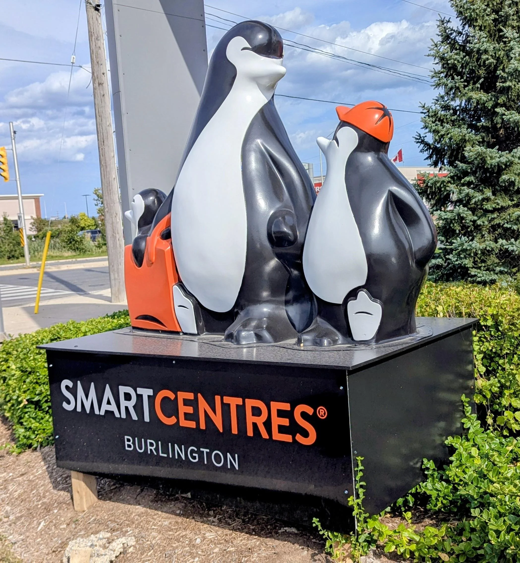 Statue of three penguins with one wearing an orange helmet next to a sign that reads 'Smart Centers Burlington.'
