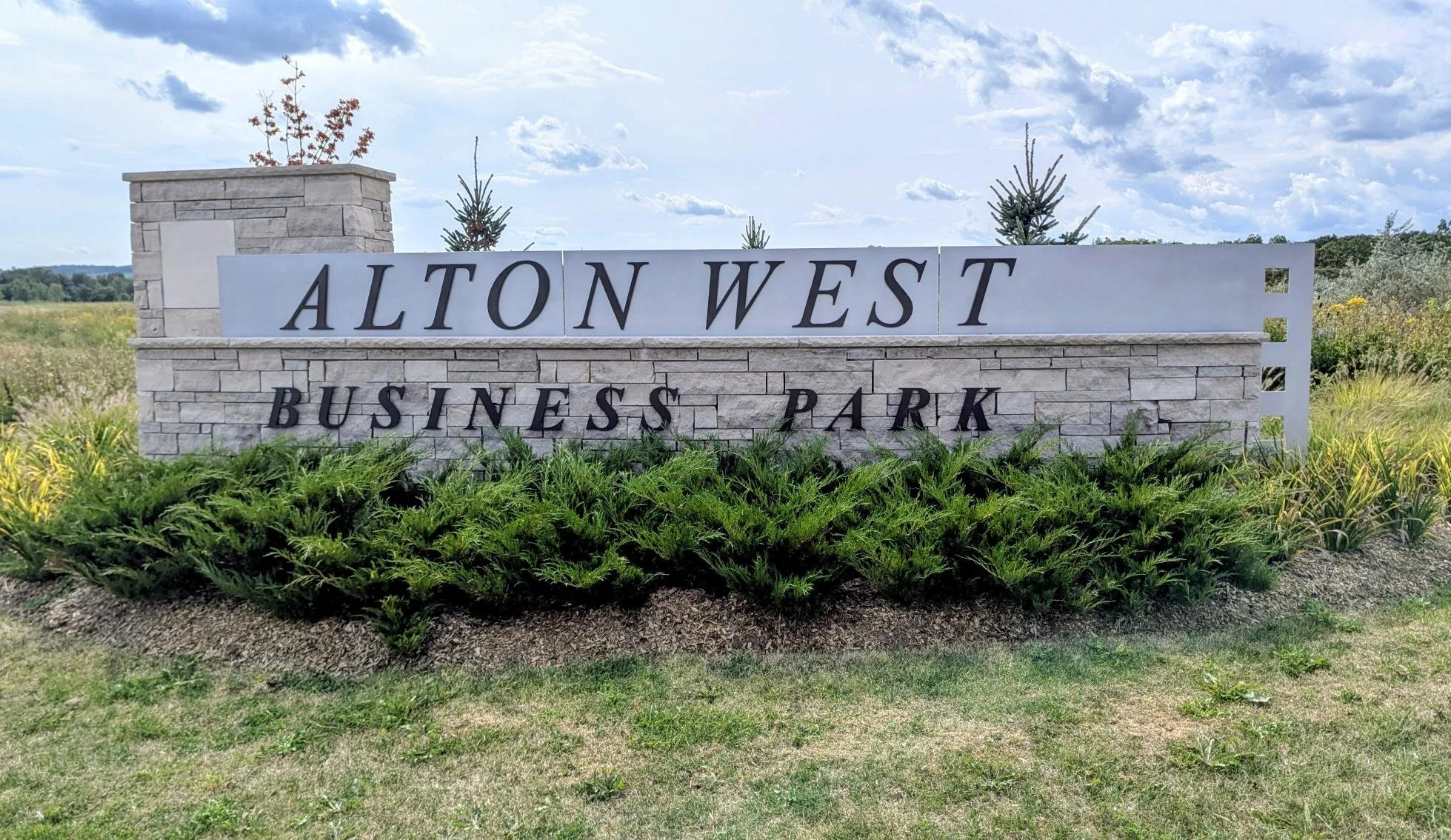 A stone and metal sign reads 'Alton West Business Park' and is surrounded by greenery and small plants, with a partly cloudy sky in the background.