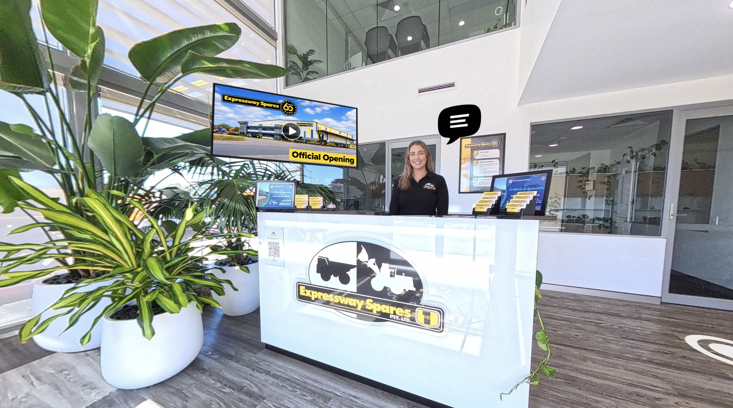 Interior of a modern office reception area with a white desk, a smiling woman in black standing behind it, large green plants in white pots, a digital screen displaying an advertisement for Expressway Spares, and glass walls with frosted graphics.