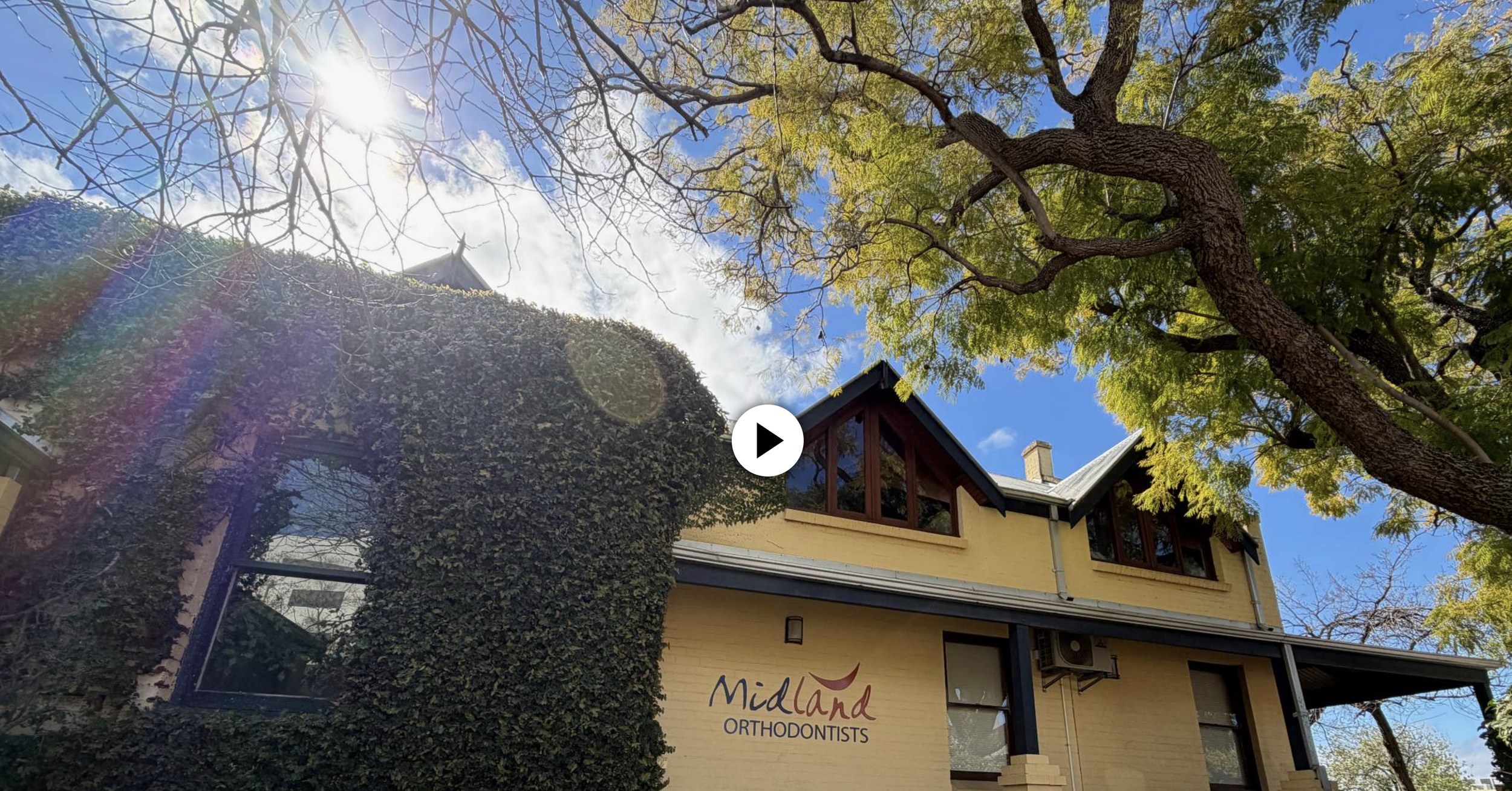 View of a house with a yellow exterior and a sign reading 'Midland Orthodontists' on the front. A large tree with green and yellow leaves is in the foreground, with the sun shining through the branches. The sky is mostly clear with some clouds.