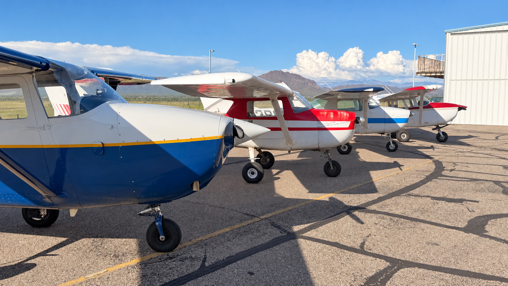 Four small airplanes parked outside near a hangar, with mountains and blue sky in the background.