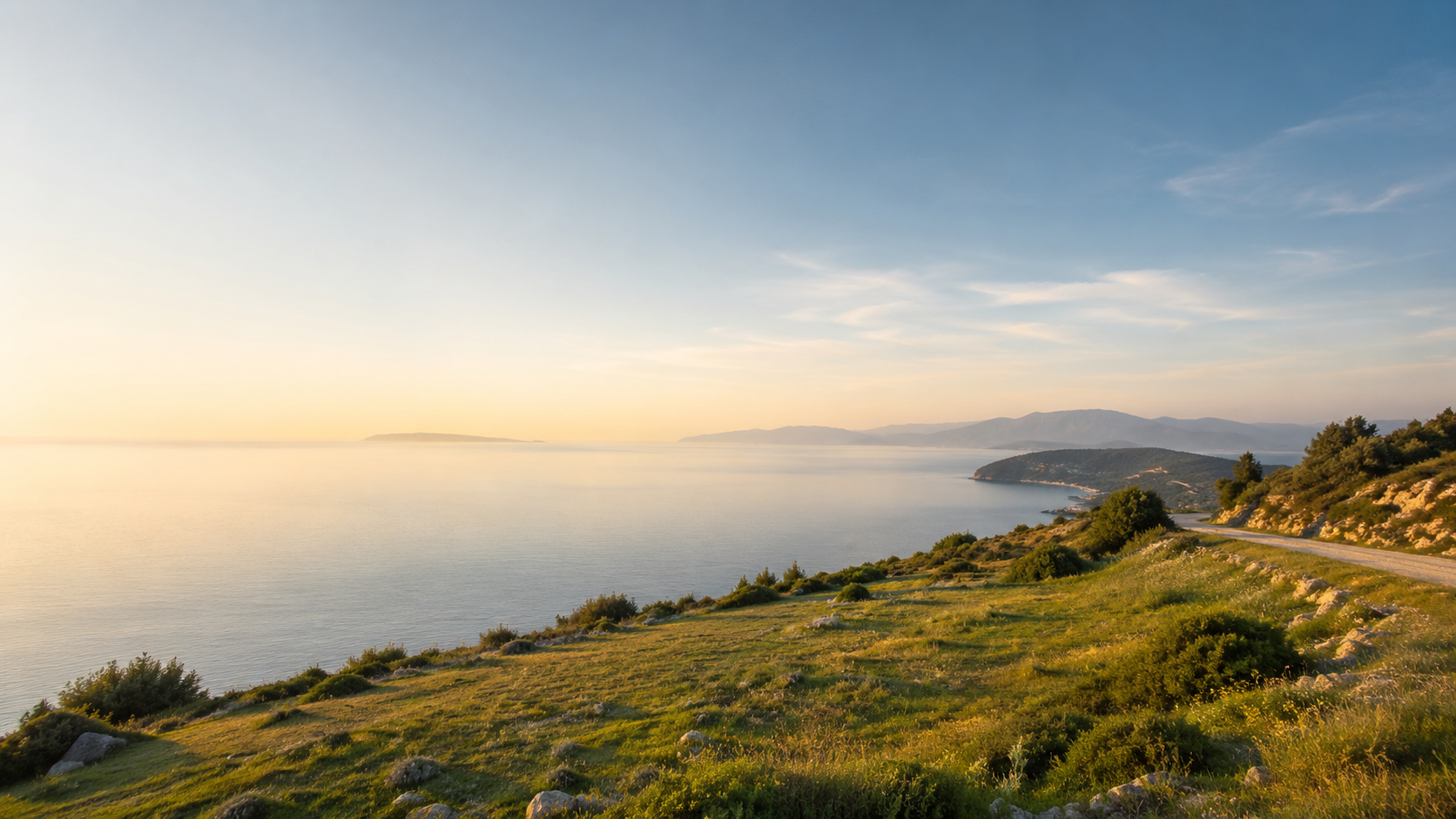Côte méditerranéenne avec mer calme, plage, collines verdoyantes et route sinueuse au coucher du soleil.