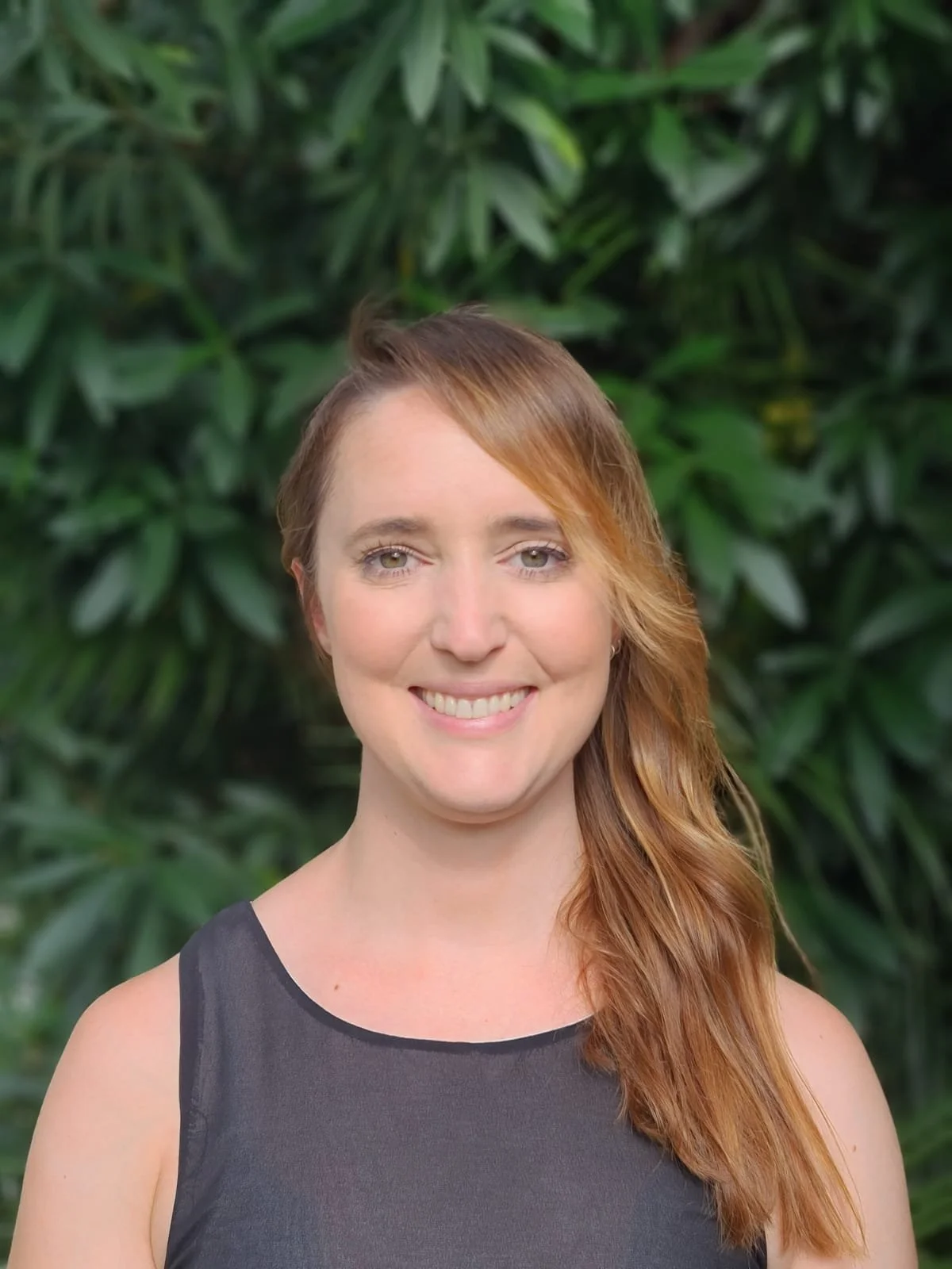 Portrait of a young woman with long red hair, smiling, wearing a sleeveless black top, standing in front of green foliage.