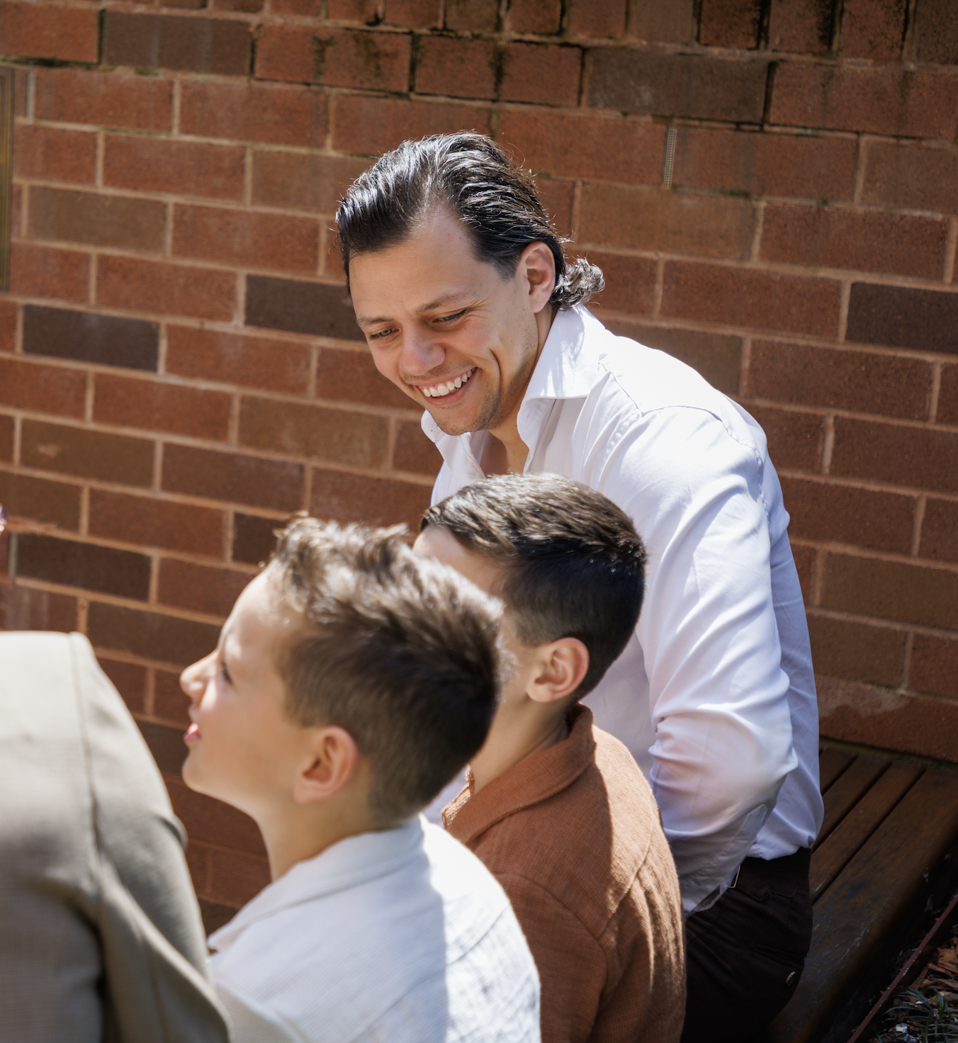 A man with dark, wavy hair and a white shirt is smiling, leaning towards two boys with short hair, who are sitting and looking up at him, against a background of a brick wall.