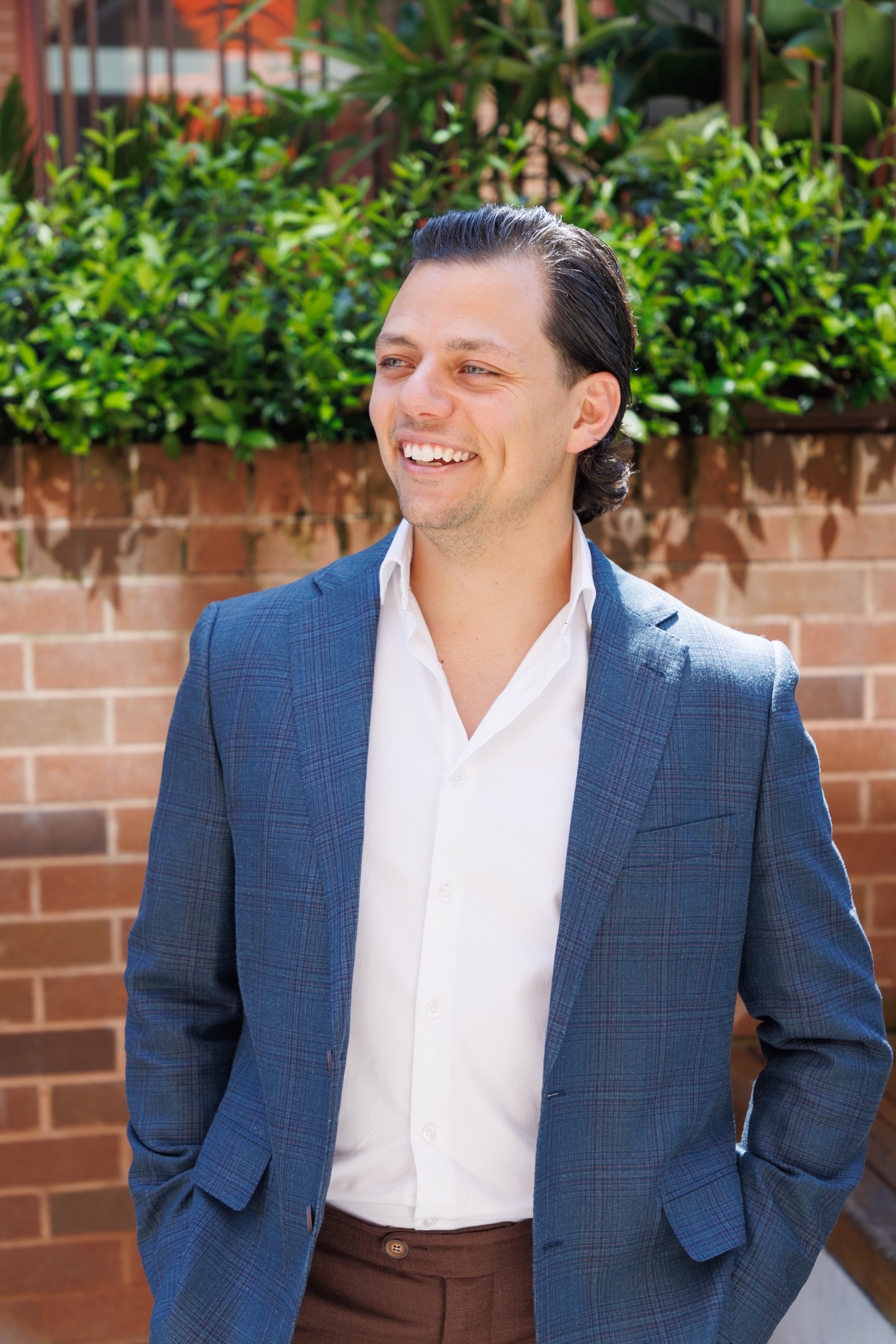 A smiling man in a blue plaid blazer and white shirt standing outdoors in front of a brick wall with green plants.