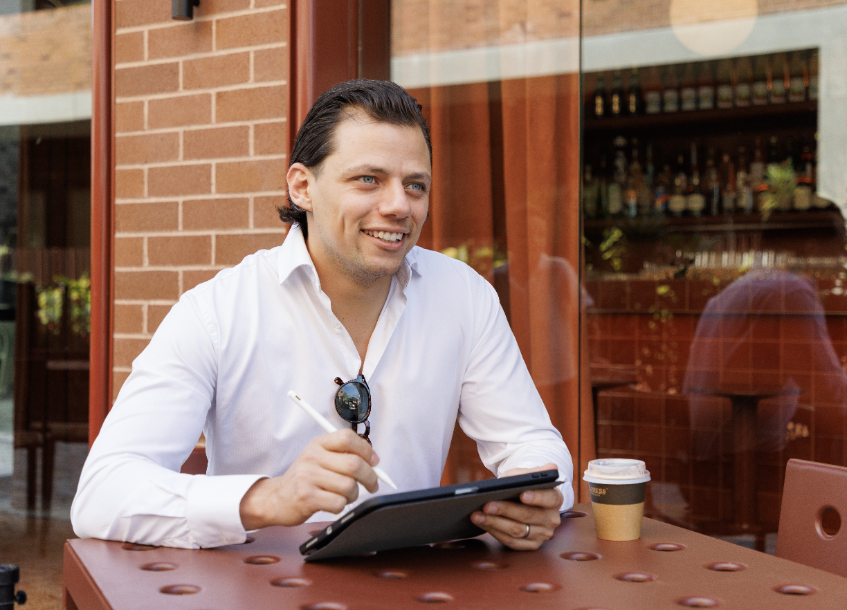 A man sitting at an outdoor cafe table using a tablet with a stylus, smiling, with sunglasses hanging on his shirt collar and a coffee cup nearby.