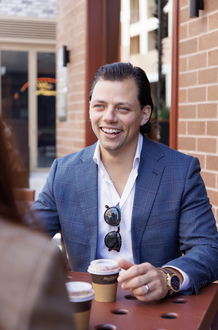 Man smiling and talking, sitting outdoors at a table with coffee cups, wearing a blue blazer, white shirt, sunglasses hanging from his shirt, and a watch on his left wrist.