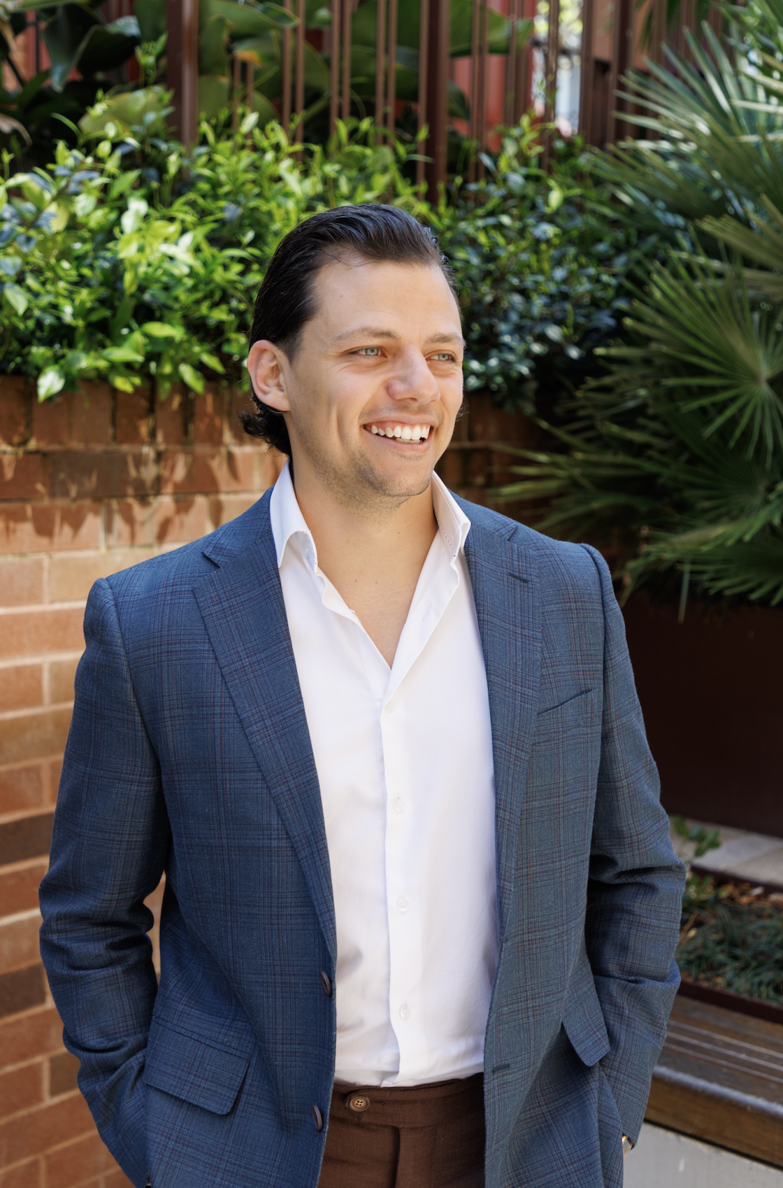 A smiling man in a blue plaid blazer and white shirt standing outdoors near a brick wall and green plants.