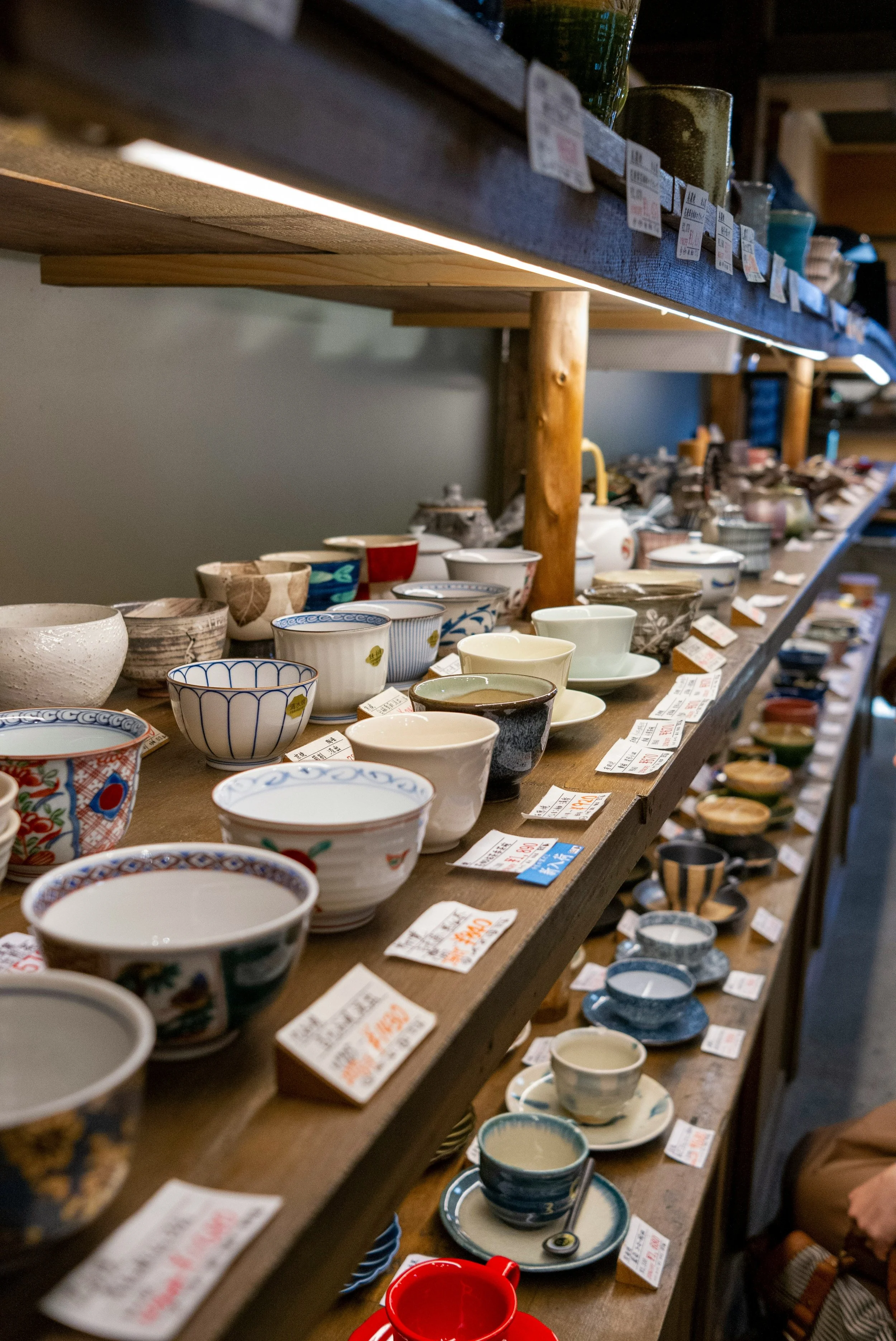 Display of various ceramic bowls and cups on wooden shelves in an antique or thrift store, with price tags attached.