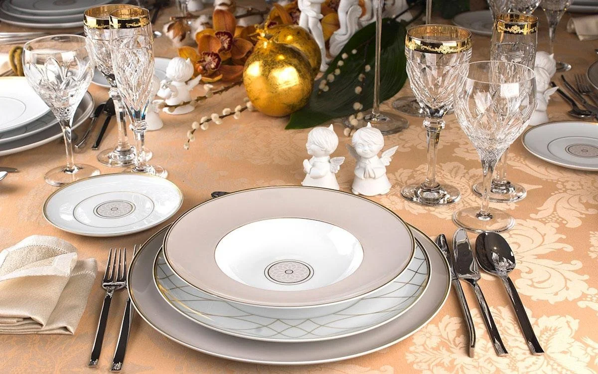 Elegant table setting with white and gold-rimmed china plates, crystal glasses, silver cutlery, and decorative angels and golden balls on a peach-colored tablecloth, with floral centerpiece and white beaded garland.