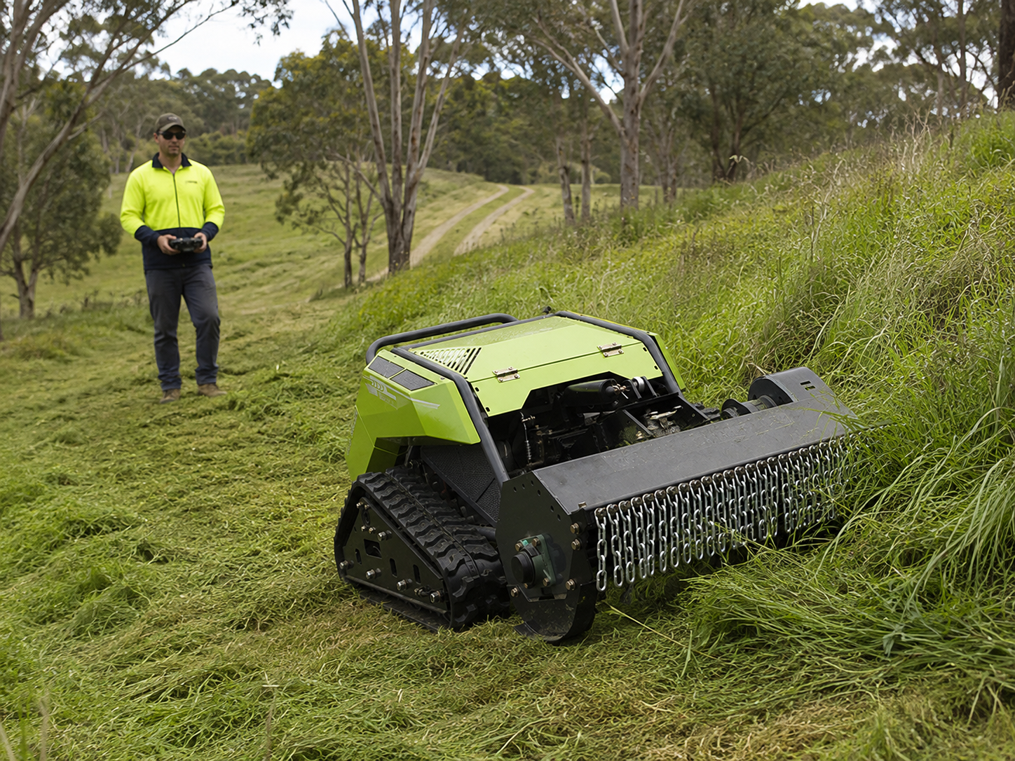 A person is operating a remote-controlled robotic lawn mower on a grassy hillside in a rural area with trees and a dirt path in the background.
