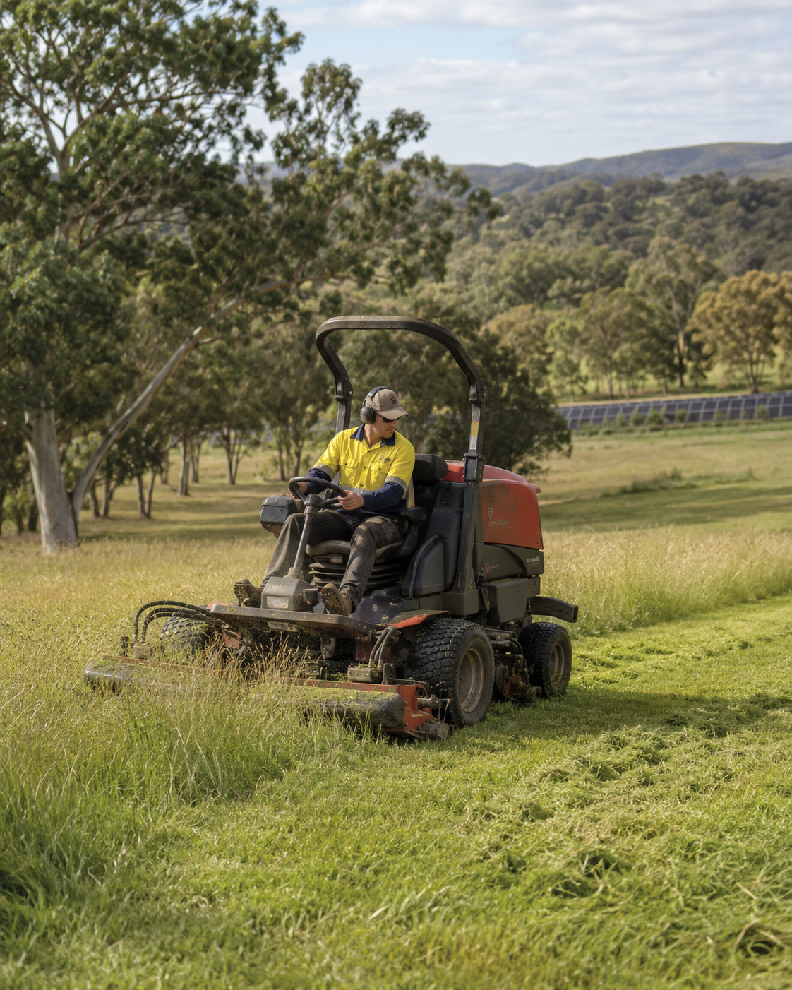 A person in safety gear riding a lawn mower through a grassy field, with trees and hills in the background.