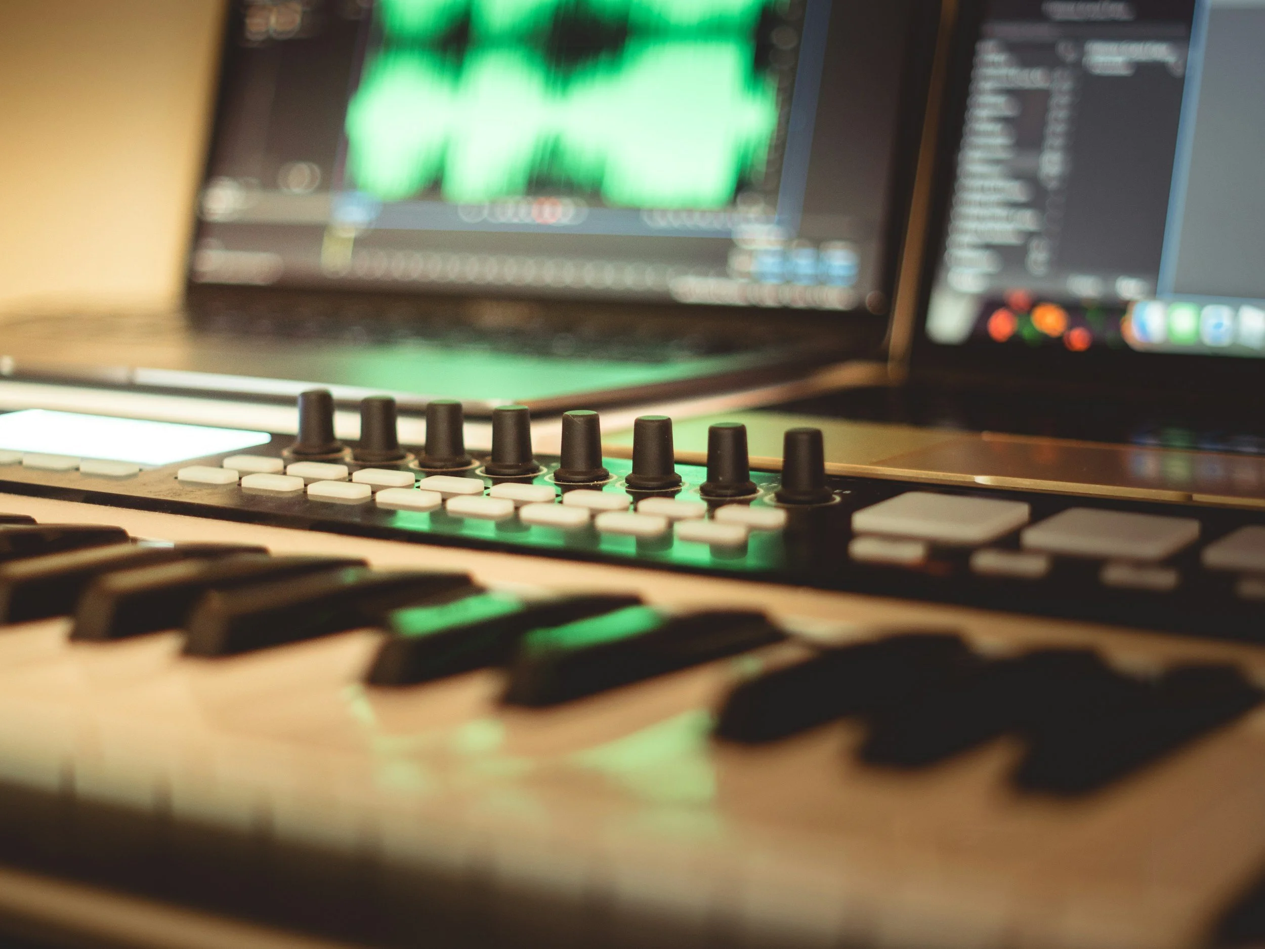 Close-up of a music production device with a computer in the background displaying a green waveform and a list of tracks.