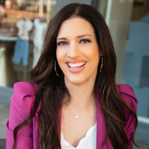 A woman with long dark hair wearing a purple blazer and smiling.