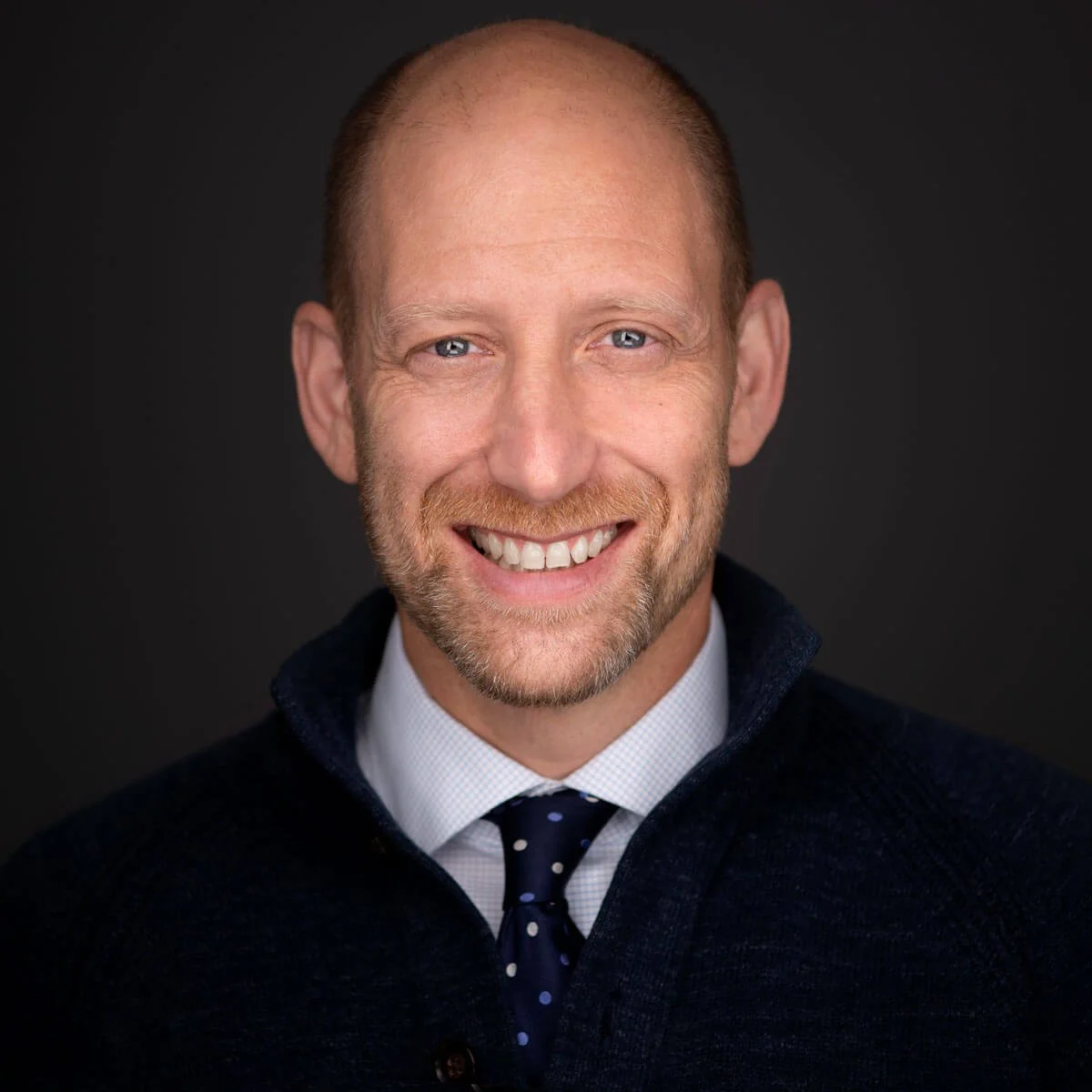 A smiling middle-aged man with a beard, wearing a dark sweater, white shirt, and a navy tie with white polka dots, set against a dark background.