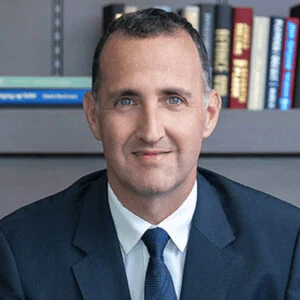 A man in a suit standing indoors with a bookshelf in the background.