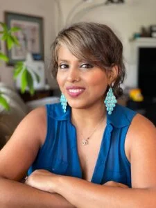 A woman with short brown hair wearing a blue sleeveless top and large dangling earrings, smiling and posing indoors.