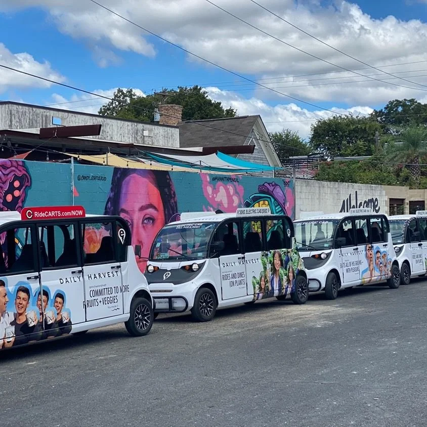 Line of Electric Cab of North America's cabs parked on a street with a colorful mural of Kittie Bubbles Cash on the wall behind them. The mural features a her face and vibrant artwork to describe her live vocals when singing.