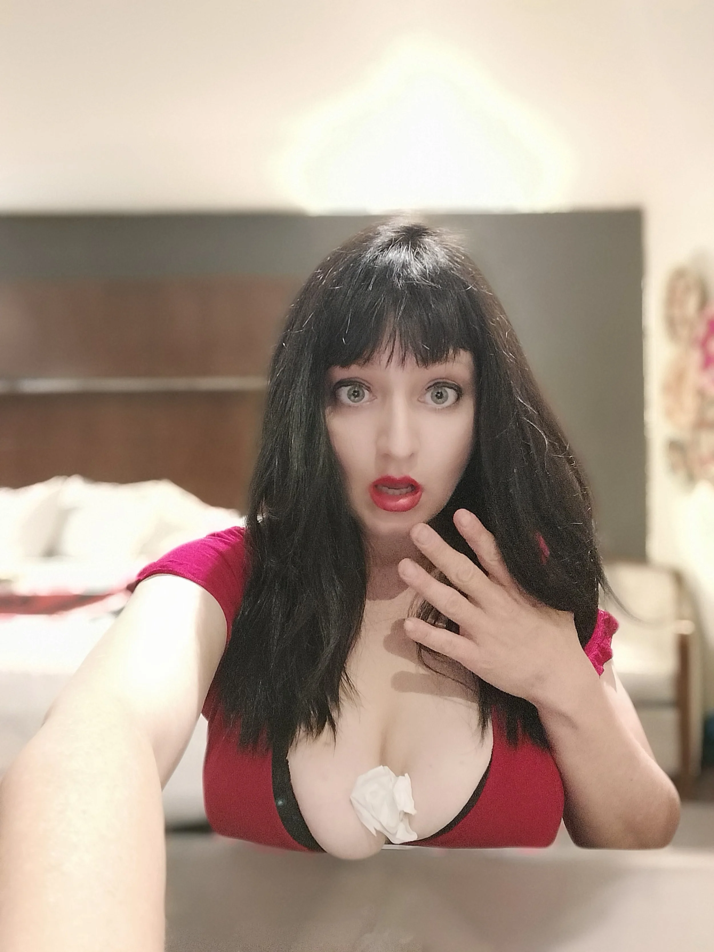 A woman with black hair and bangs taking a selfie in front of a mirror. She is wearing a red top with black trim and has red lipstick. Her left hand is near her face, and she is holding the camera with her right hand. The background shows a blurred bedroom with bed and furniture.