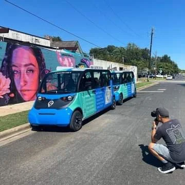 A person crouching and taking a photo of a small blue and black autonomous bus on a street with a mural of Kittie Bubbles Cash on the wall behind.