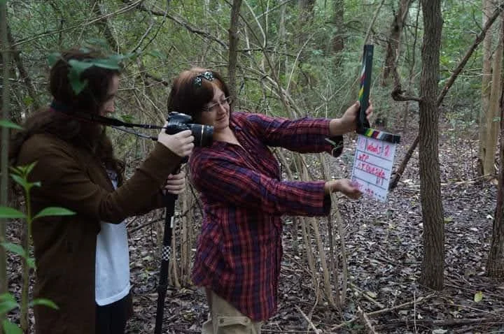 Kittie Bubbles Cash in a forest setting, one holding a camera and the other holding a clapboard, appearing to film.