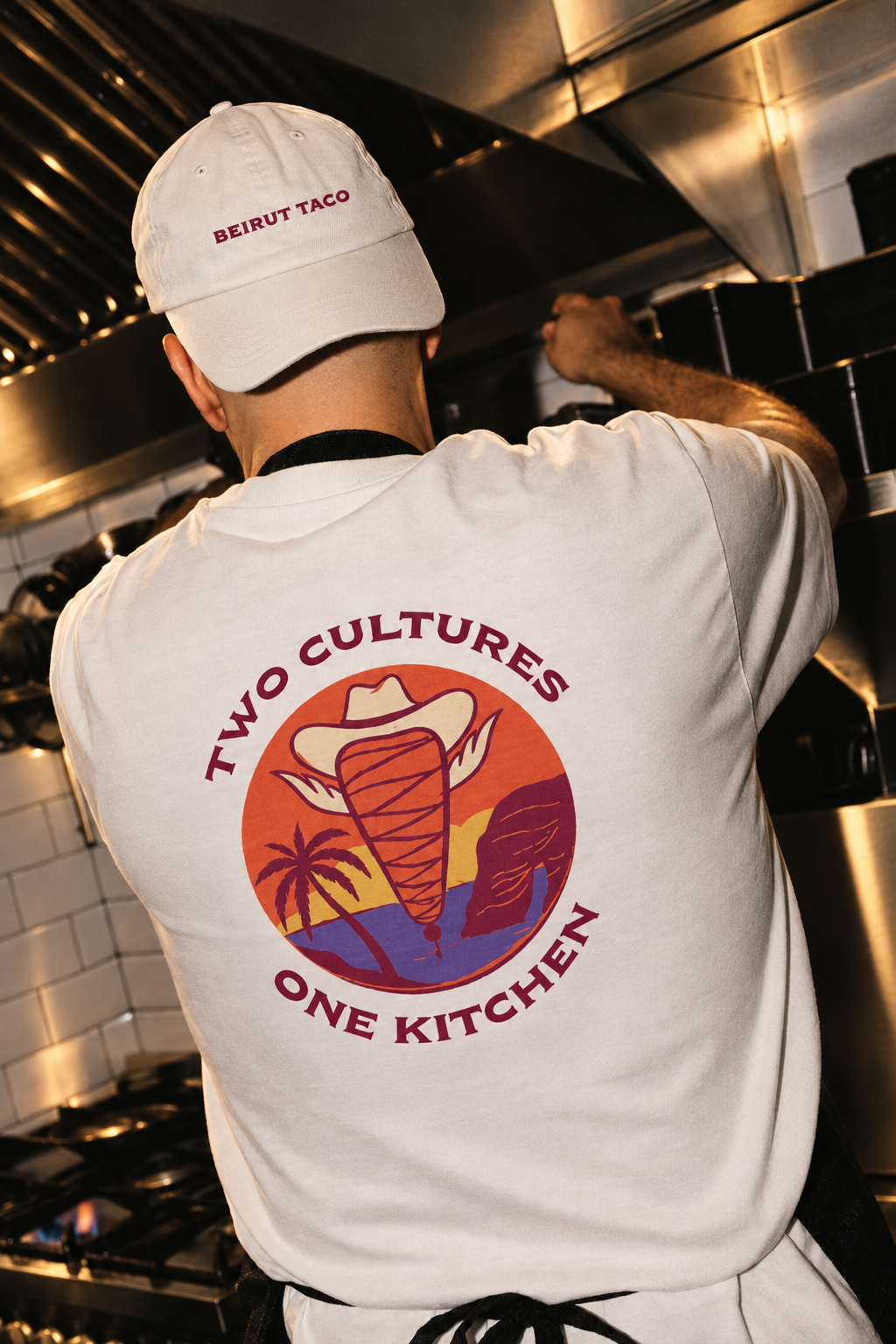 Back view of a person in a white cap and apron cooking in a kitchen with a logo that reads 'Two Cultures, One Kitchen' featuring a hat, palm trees, a mountain, and a seaside sunset.